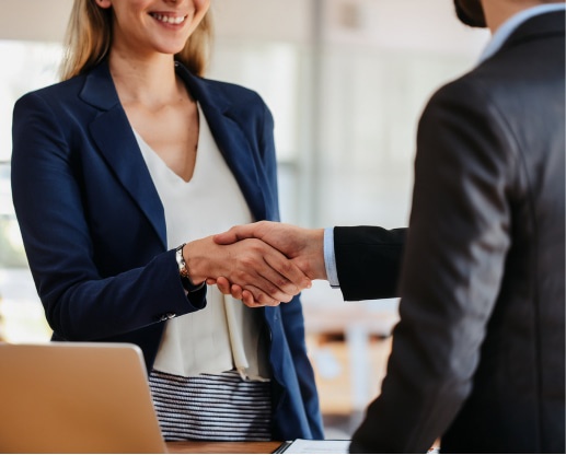 Esecuzioni Mobiliari e Immobiliari. Due professionisti si stringono la mano in un ambiente aziendale moderno. La donna indossa un blazer blu, l'uomo è in giacca formale. Sullo sfondo si intravede un laptop e un ambiente luminoso.